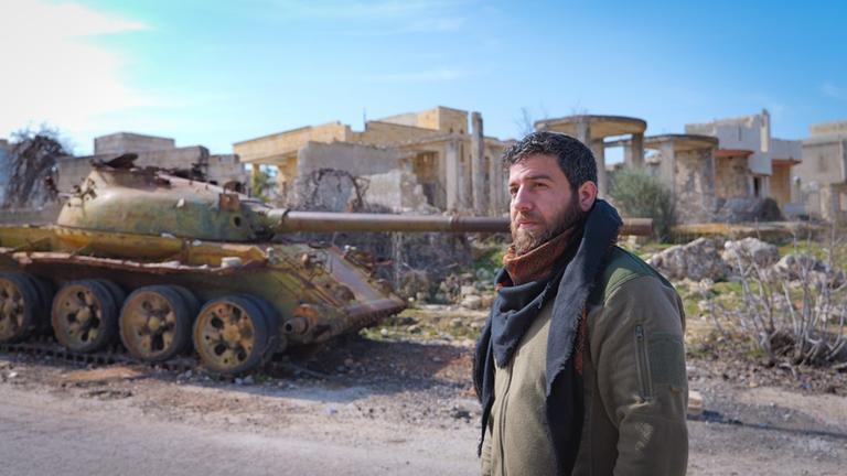 Mustafa Al-Aloush steht auf der Straße, im Hintergrund ein Panzer und zerstörte Gebäude