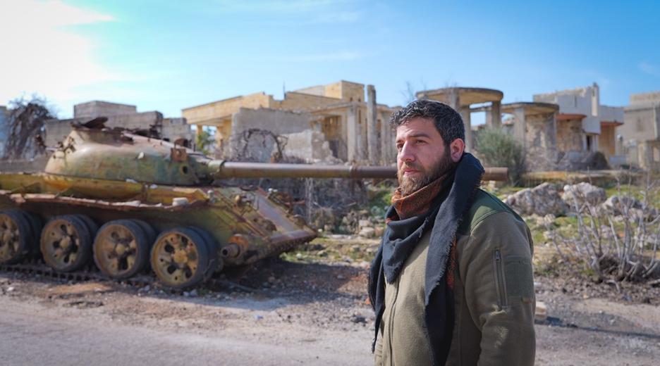 Mustafa Al-Aloush steht auf der Straße, im Hintergrund ein Panzer und zerstörte Gebäude
