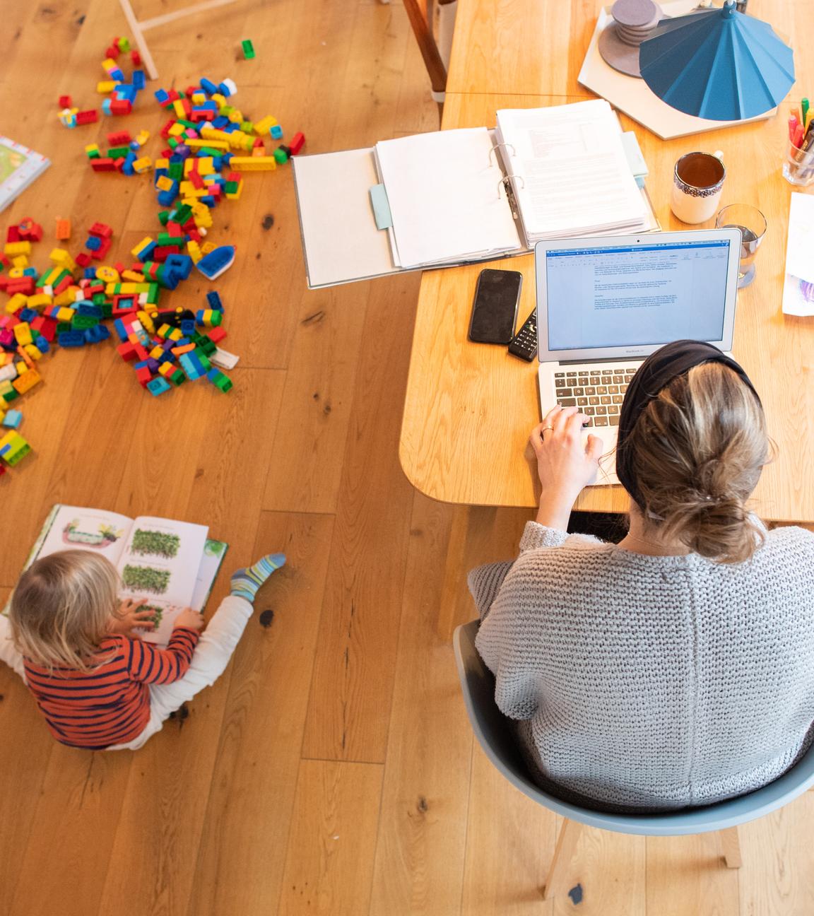 Ein zweijähriges Kind spielt im Wohnzimmer, während seine Mutter Zuhause im Homeoffice an einem Laptop arbeitet.