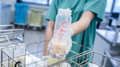 Flaschen mit Muttermilch in Drahtkörben in einer Frauenmilchbank. Darüber hält eine Hand eine Flasche mit gespendeter Muttermilch.