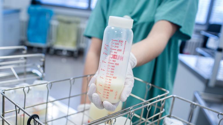 Flaschen mit Muttermilch in Drahtkörben in einer Frauenmilchbank. Darüber hält eine Hand eine Flasche mit gespendeter Muttermilch.