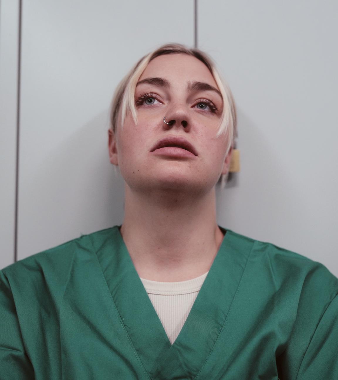 Eine junge Frau trägt einen grünen Kasack und lehnt an der Wand. Sie schaut nach oben und sieht erschöpft aus.
