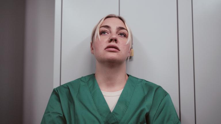 Eine junge Frau trägt einen grünen Kasack und lehnt an der Wand. Sie schaut nach oben und sieht erschöpft aus.