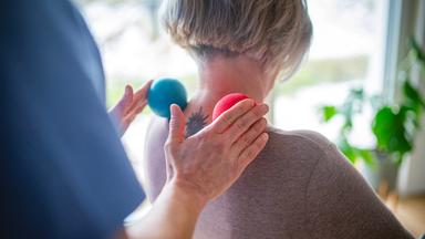 Physiotherapeutin hilft einer älteren Frau bei einer Nackenübung mit Massagebällen.