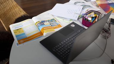 Ein Laptop steht aufgeklappt auf dem Tisch und davor liegen viele Bücher und Hefte.