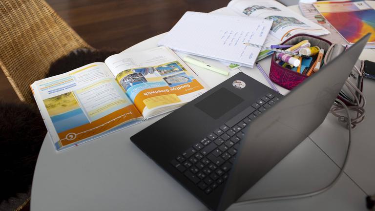 Ein Laptop steht aufgeklappt auf dem Tisch und davor liegen viele Bücher und Hefte.