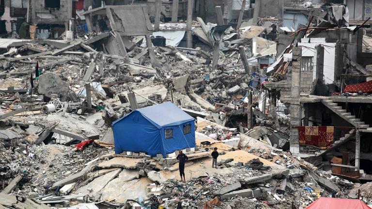 Menschen stehen am 08.04.2025 vor ihrem Zelt auf den Trümmern eines zerstörten Gebäudes im Lager Jabalia 