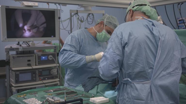 Zwei Chirurgen stehen an einem OP-Tisch und operieren eine verkrümmte Nasenscheidewand. Im Hintergrund ist die Operation an der Nase auf einem Monitor zu sehen.