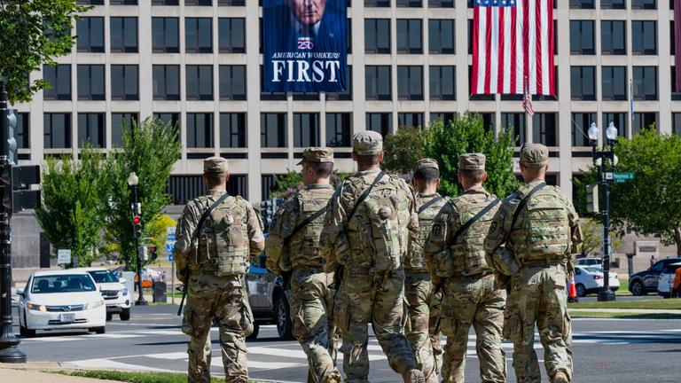 26.08.2025, USA, Washington: Bewaffnete Soldaten der Nationalgarde aus West Virginia patrouillieren auf der Mall in der Nähe des Arbeitsministeriums, wo ein Banner von US-Präsident Trump hängt.