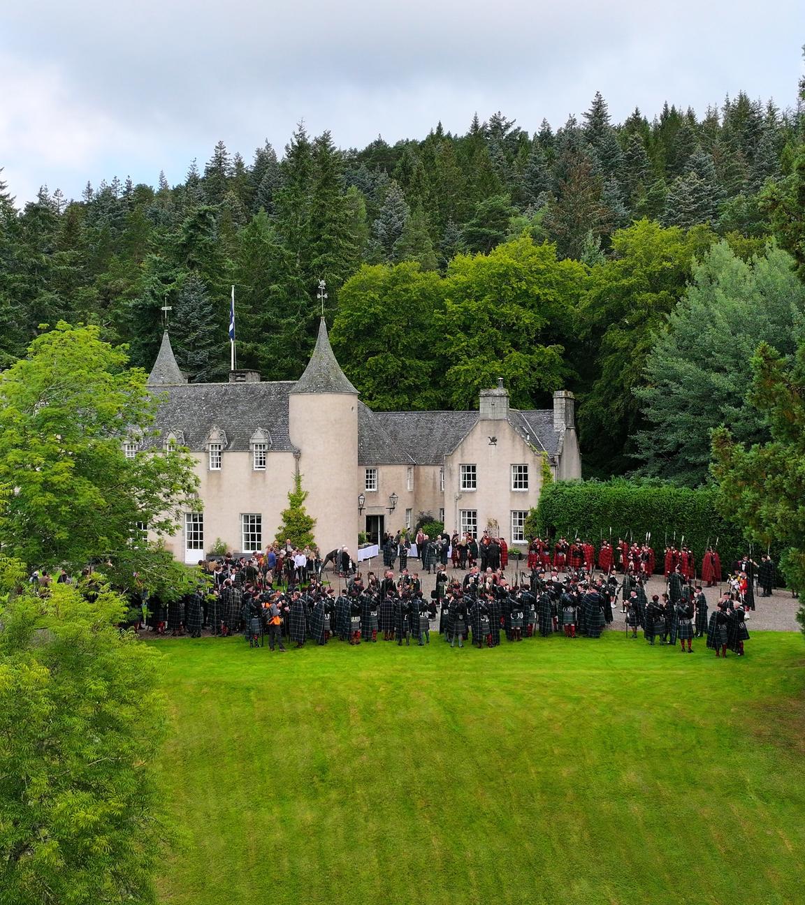  Blick von schräg oben: Vor einem schlossartigen Gebäude in grüner Landschaft stehen Menschen.