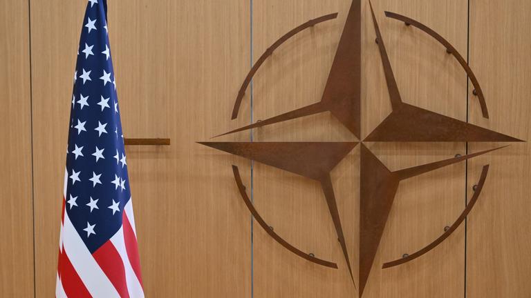 Eine amerikanische Flagge steht links. Das Emblem der Nato ist an der hölzernen Wand zu sehen.