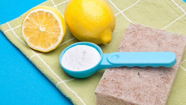 Ein Dosierlöffel weißes Natronpulver, ein Schwamm, eine ganze und eine halbe Zitrone liegen auf einem Geschirrtuch.