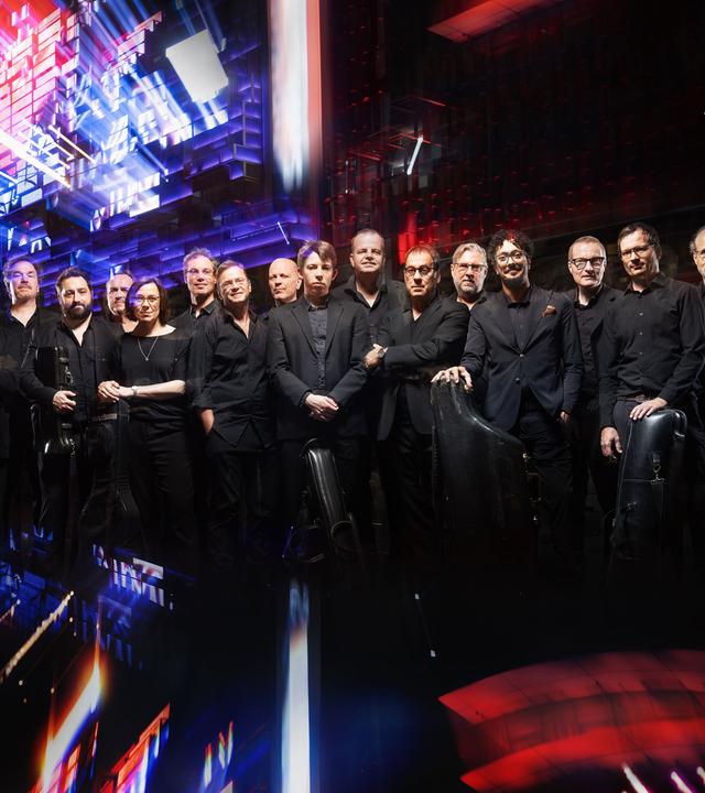 Große Musikgruppe auf einer Bühne mit bunten Lichteffekten; mehrere Musiker mit Instrumenten in Konzertaufstellung.