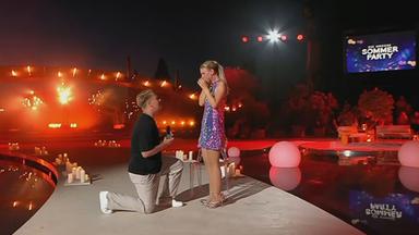 Auf dem Bild zu sehen: Ein Mann der sich vor einer Frau auf einer Bühne hinkniet und einen Heiratsantrag macht.