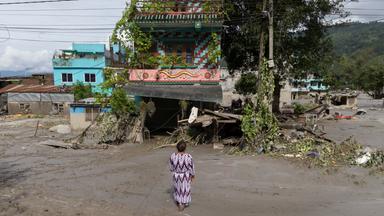 Eine Frau steht auf einer von Schlamm überspülten Straße vor ihrem beschädigten Haus in Nepal.