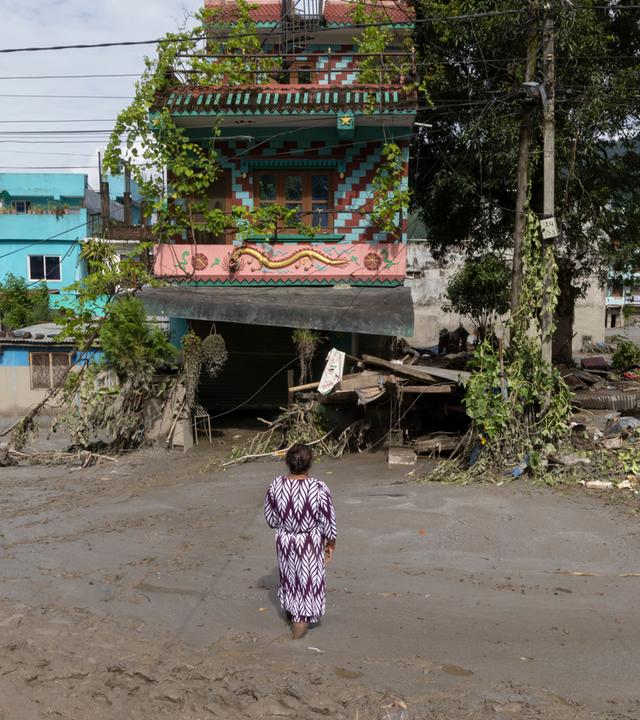 Eine Frau steht auf einer von Schlamm überspülten Straße vor ihrem beschädigten Haus in Nepal.