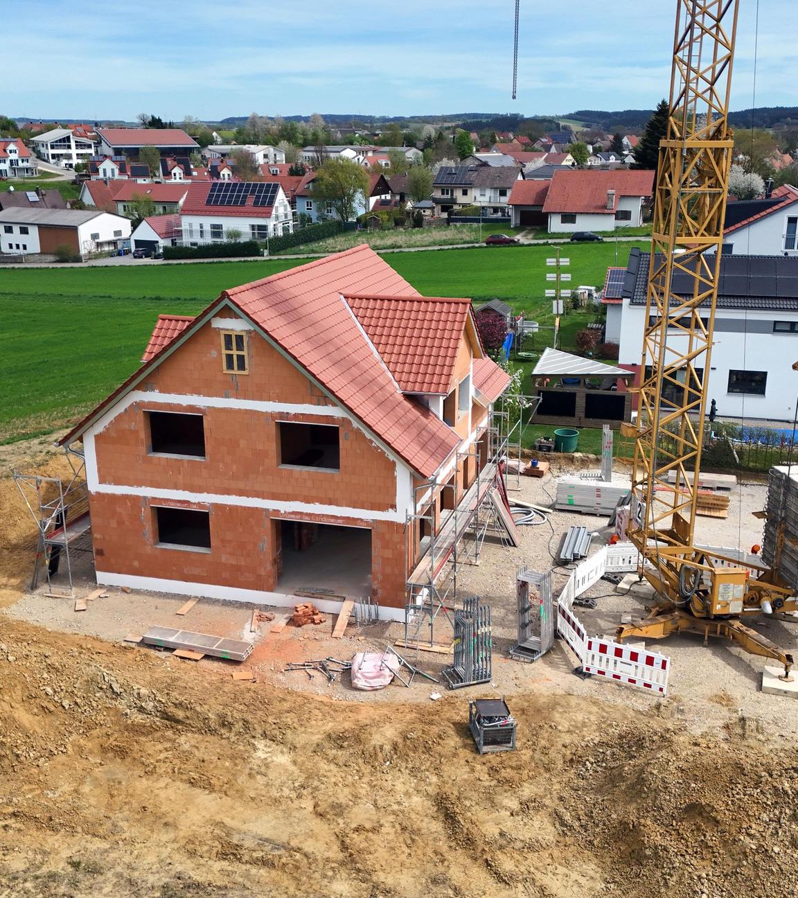 Baustelle mit dem Rohbau eines Einfamilienhauses aus rotem Backstein und mit rotem Ziegeldach. Drumherum aufgeschütteter Sand und ein Kran. Im Hintergrund ein Feld und die anderen Häuser des Dorfes.