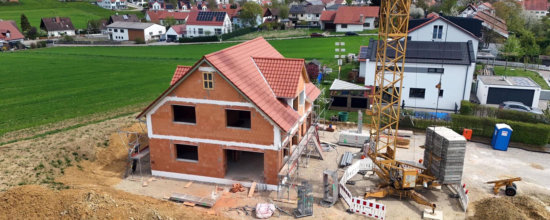 Baustelle mit dem Rohbau eines Einfamilienhauses aus rotem Backstein und mit rotem Ziegeldach. Drumherum aufgeschütteter Sand und ein Kran. Im Hintergrund ein Feld und die anderen Häuser des Dorfes.