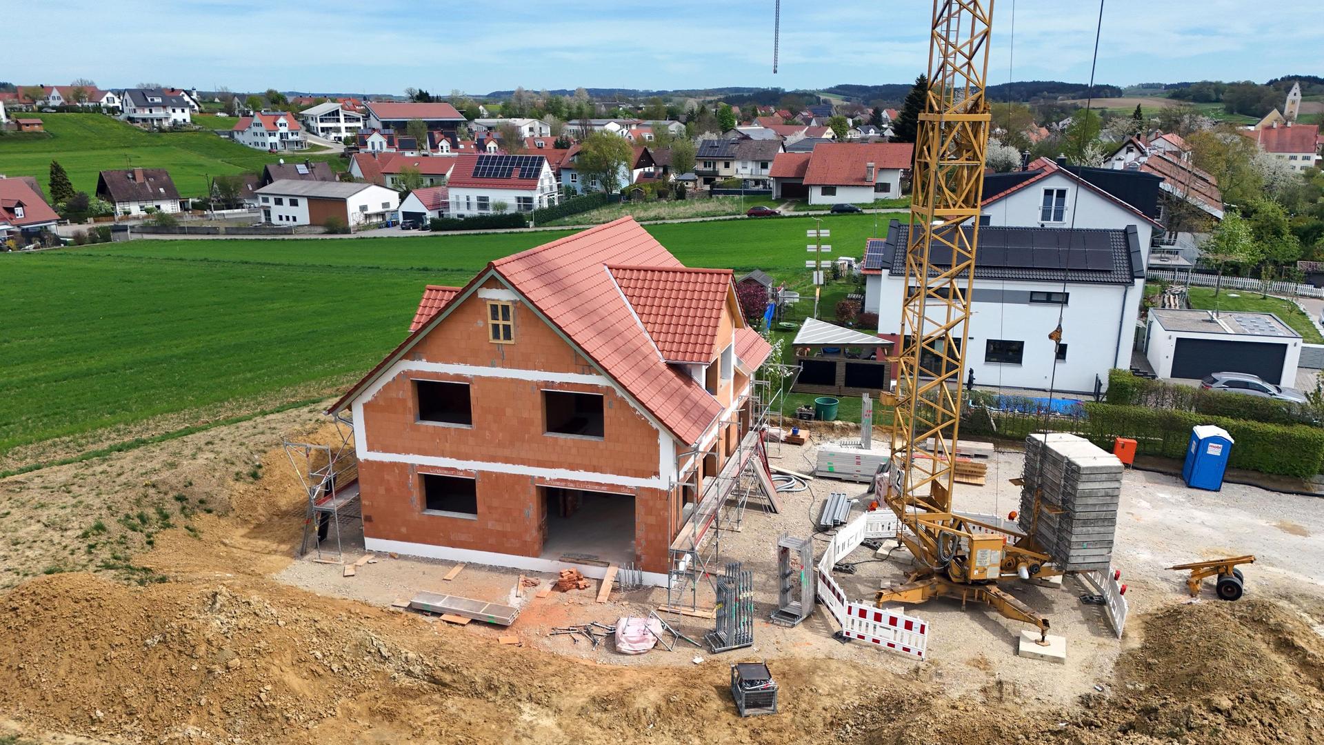 Baustelle mit dem Rohbau eines Einfamilienhauses aus rotem Backstein und mit rotem Ziegeldach. Drumherum aufgeschütteter Sand und ein Kran. Im Hintergrund ein Feld und die anderen Häuser des Dorfes.