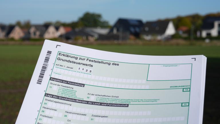 Ein Erklärung zur Feststellung des Grundsteuerwerts für das Jahr 2025 wird vor eine Wohnsiedlung hochgehalten.