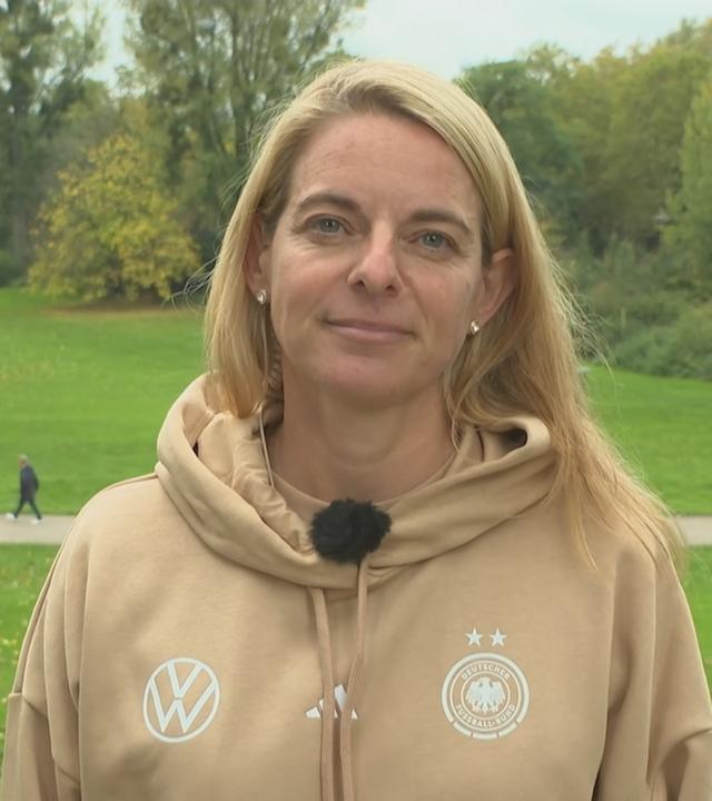 DFB-Sportdirektorin Nia Künzer am ZDF-Mikrofon. Im Hintergrund ist ein Park zu sehen mit viel Grün.