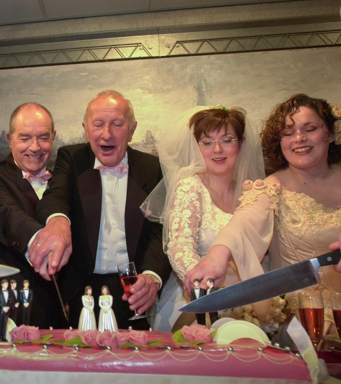 Sechs Personen in schwarzen Anzügen und weißen Hochzeitskleidern schneiden eine Torte an.