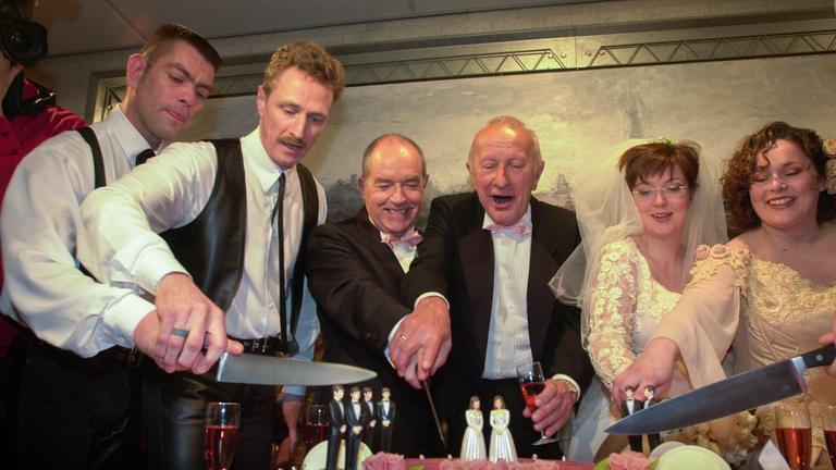 Sechs Personen in schwarzen Anzügen und weißen Hochzeitskleidern schneiden eine Torte an.