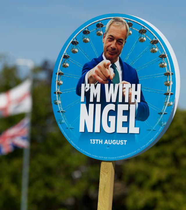 Ein rundes Schild zur Wahlwerbung mit dem Gesich von Nigel Farage, dahinter sind die Fahnen Englands und Großbritanniens zu sehen.
