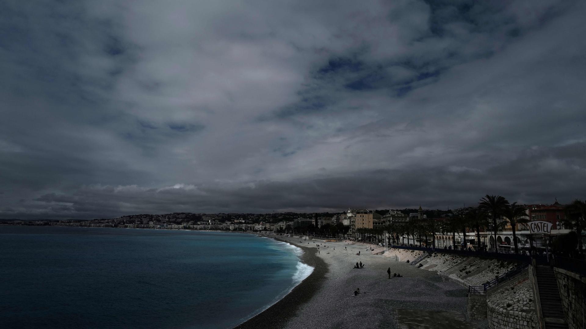 Menschen sitzen am Strand oder spazieren entlang der berühmten Promenade des Anglais unter einem bewölkten Himmel in Nizza.