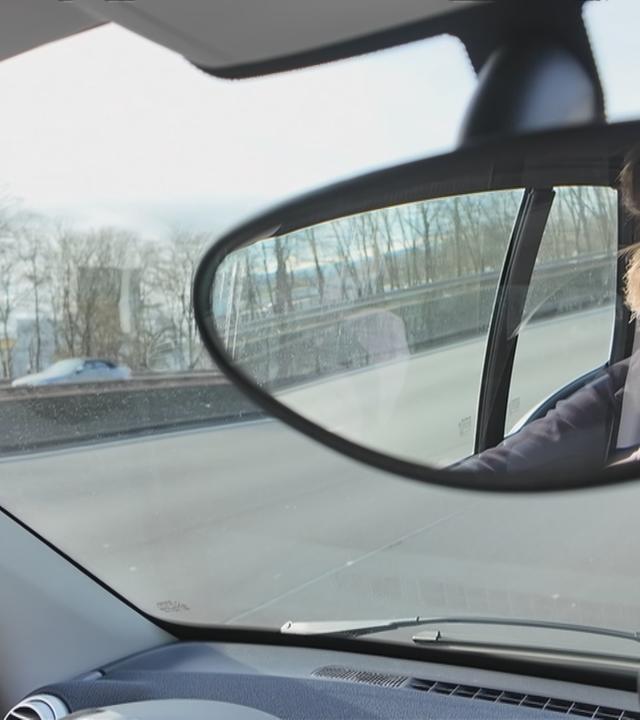 Frau fährt Auto, man sieht ihr Gesicht im Innenspiegel des Autos. 