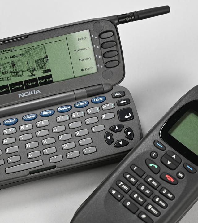 Zwei Nokia 9000 Communicator, einmal aufgeklappt, mit sichtbarem Display und Tastatur, darunter einmal zugeklappt, liegen auf einem hellen Hintergrund. 