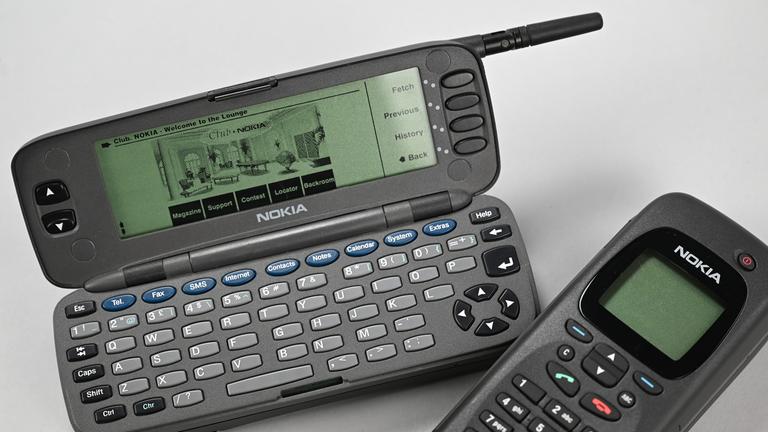 Zwei Nokia 9000 Communicator, einmal aufgeklappt, mit sichtbarem Display und Tastatur, darunter einmal zugeklappt, liegen auf einem hellen Hintergrund. 