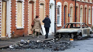 Menschen gehen an den Überresten eines ausgebrannten Autos vorbei, das vor vernagelten Häusern nahe der Newtownards Road im Osten von Belfast (Nordirland) steht.