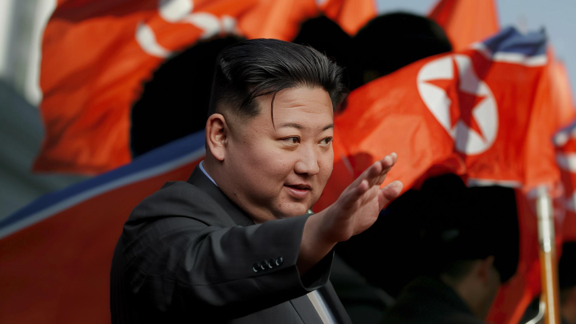 Kim Jong Un trägt einen schwarzen Anzug und streckt den rechten Arm nach vorne. Im Hintergrund wehen nordkoreanische Flaggen.