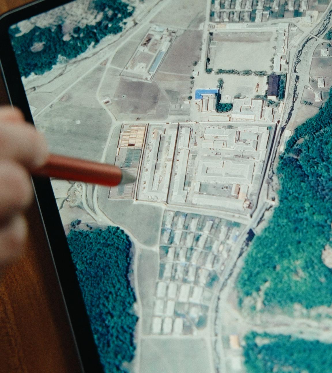 Eine Hand zeigt mit einem Stift auf ein Computerdisplay. Darauf ist ein Satellitenbild mit einer größeren Anlage neben einem Wald in Nordkorea zu erkennen.