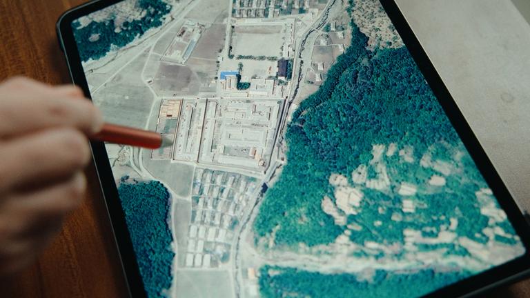 Eine Hand zeigt mit einem Stift auf ein Computerdisplay. Darauf ist ein Satellitenbild mit einer größeren Anlage neben einem Wald in Nordkorea zu erkennen.