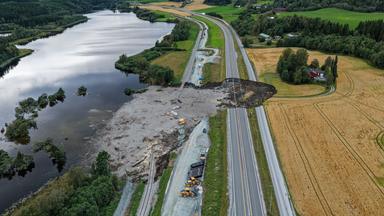 Von einem großen Loch in einer Landstraße aus fließt schlamm in einen anliegenden See.