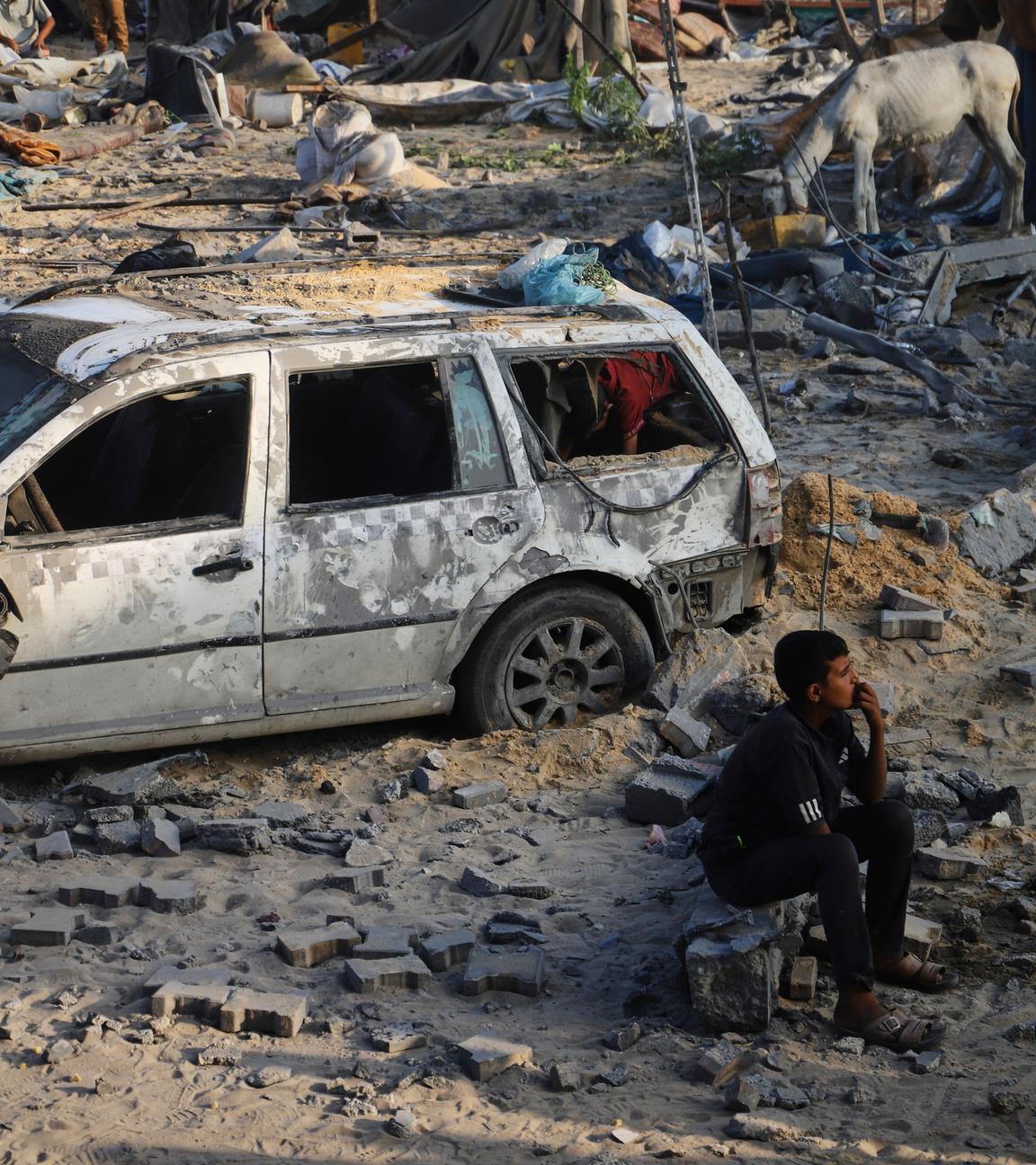 Ein Junge sitzt vor einem kaputten Auto im Sand im Gazastreifen.