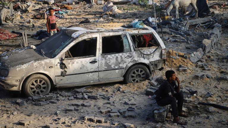 Ein Junge sitzt vor einem kaputten Auto im Sand im Gazastreifen.