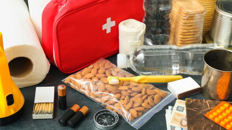 Utensilien wie Taschenlampe, Wasserflaschen, Erste-Hilfe-Set, Reisepass, Bargeld und Streichhölzer für einen Notfallrucksack auf einem Tisch.