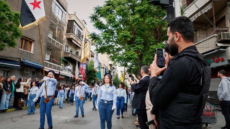  Ein Sicherheitsbeamter filmt mit seinem Smartphone, wie Mitglieder der griechisch-orthodoxen Kirche am Palmsonntag durch das überwiegend christliche Viertel al-Qassaa ziehen. Am 13.04.2025, in Syrien, Damaskus