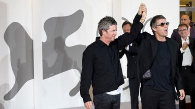 Zwei schwarz gekleidete Männer sthen vor einer weißen Wand mit dem grauen Löwen-Logo der Filmfestspiele von Venedig, halten sich an einer Hand und heben diese hoch.