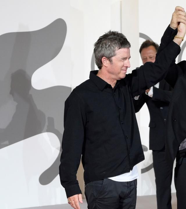 Zwei schwarz gekleidete Männer sthen vor einer weißen Wand mit dem grauen Löwen-Logo der Filmfestspiele von Venedig, halten sich an einer Hand und heben diese hoch.