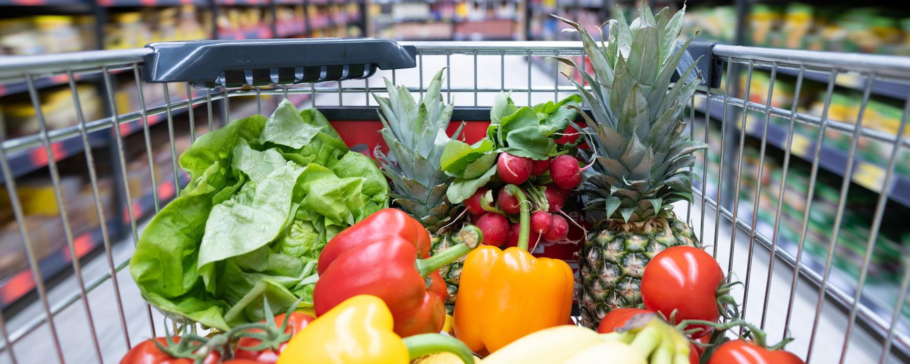 Ein Einkaufskorb mit Obst und Gemüse steht in einem Supermarkt.