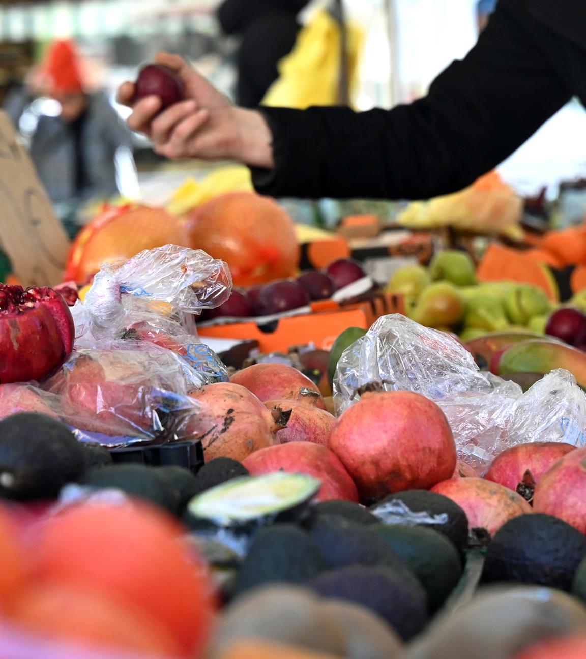 Eine Person hält an einem Obststand am Marktplatz in Leipzig eine Pflaume und eine Einkaufstüte in der Hand