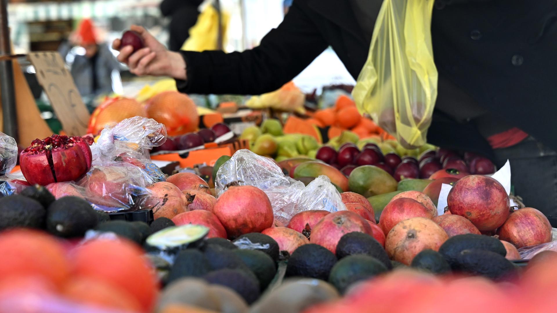 Eine Person hält an einem Obststand am Marktplatz in Leipzig eine Pflaume und eine Einkaufstüte in der Hand