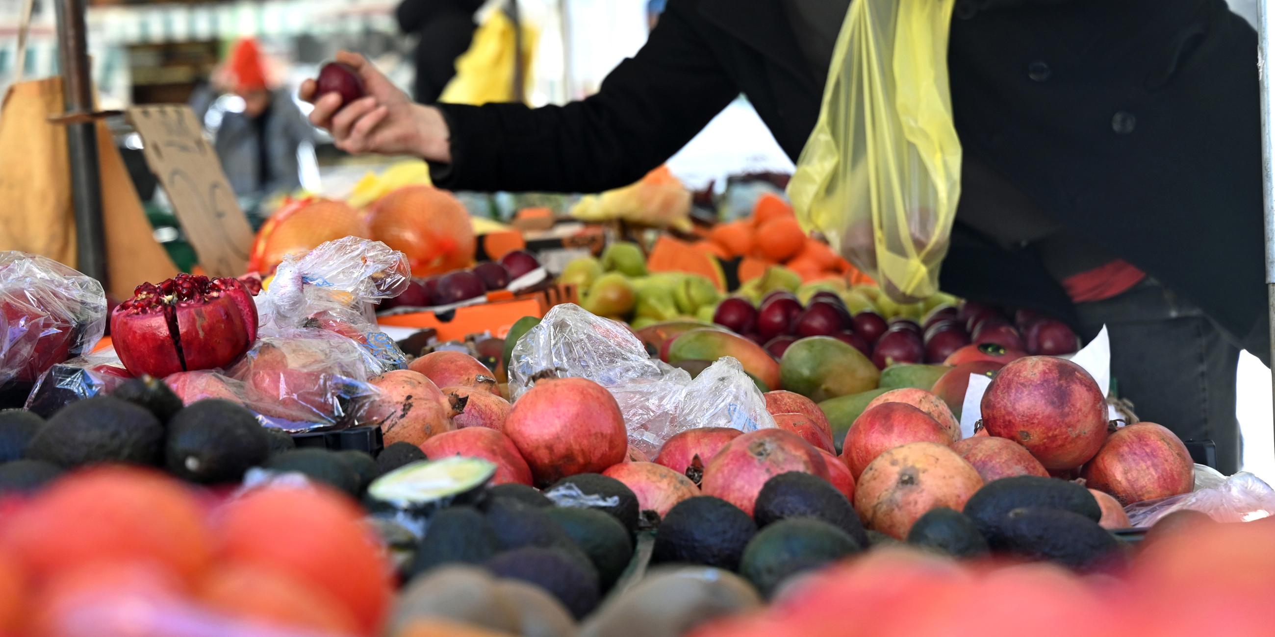 Eine Person hält an einem Obststand am Marktplatz in Leipzig eine Pflaume und eine Einkaufstüte in der Hand