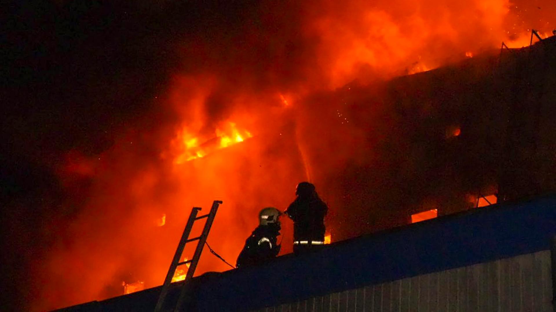 Ein Wohnhaus steht nachts in Rauch und Flammen, zwei Feuerwehrleute stehen davor.