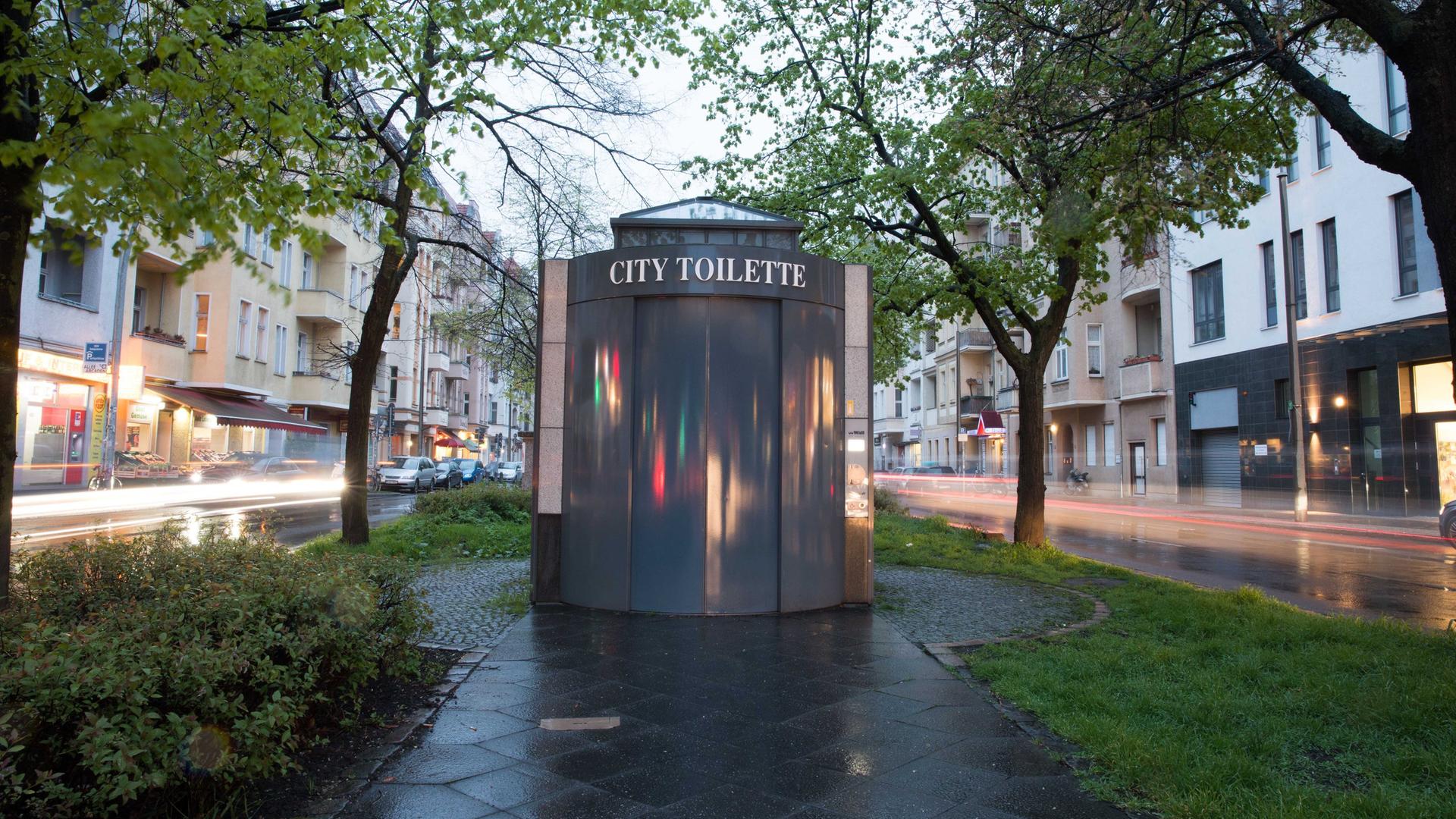 Eine öffentliche Toilette steht auf einem Grünstreifen mit Gehweg zwischen zwei Straßen.