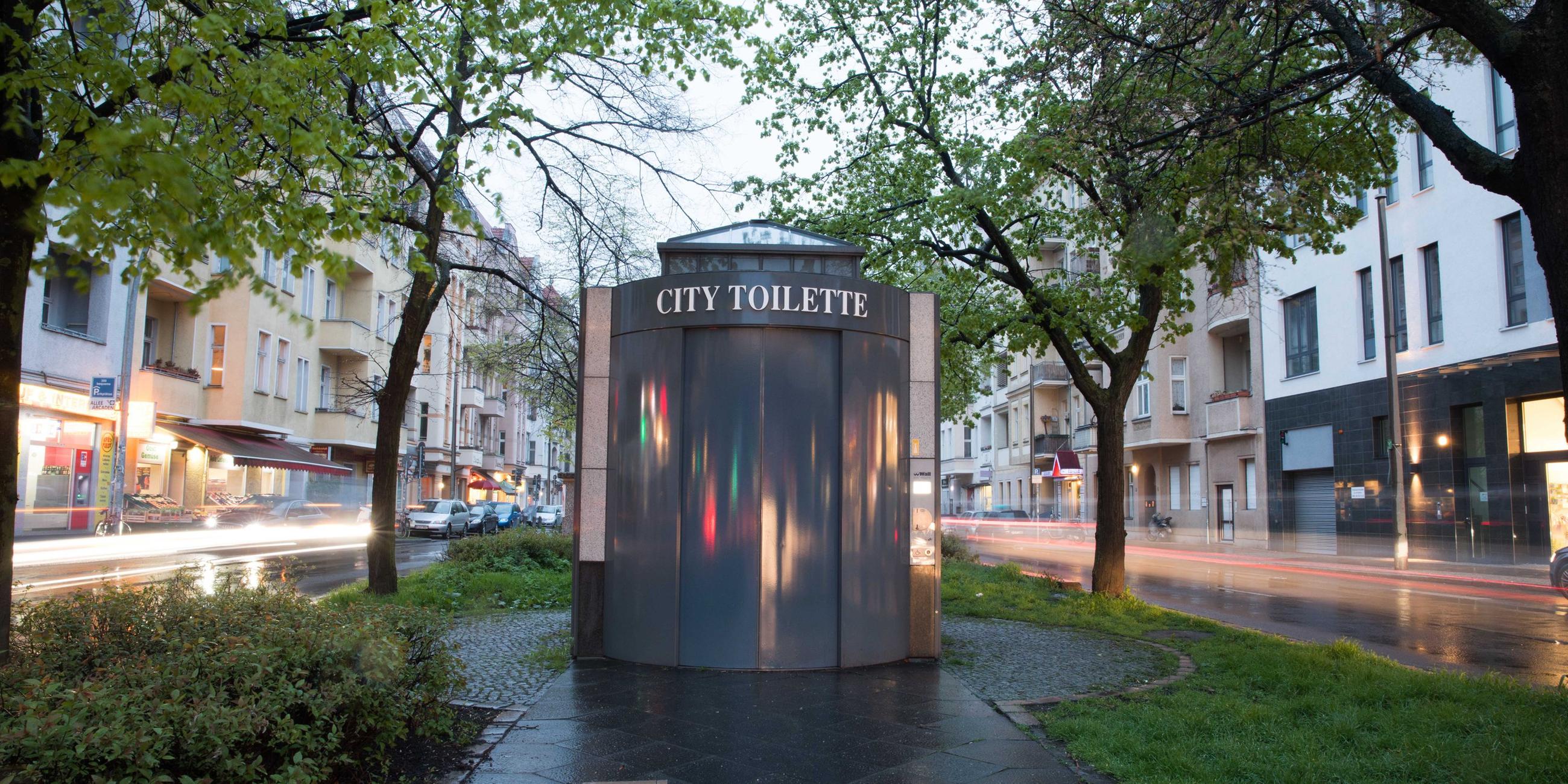 Eine öffentliche Toilette steht auf einem Grünstreifen mit Gehweg zwischen zwei Straßen.